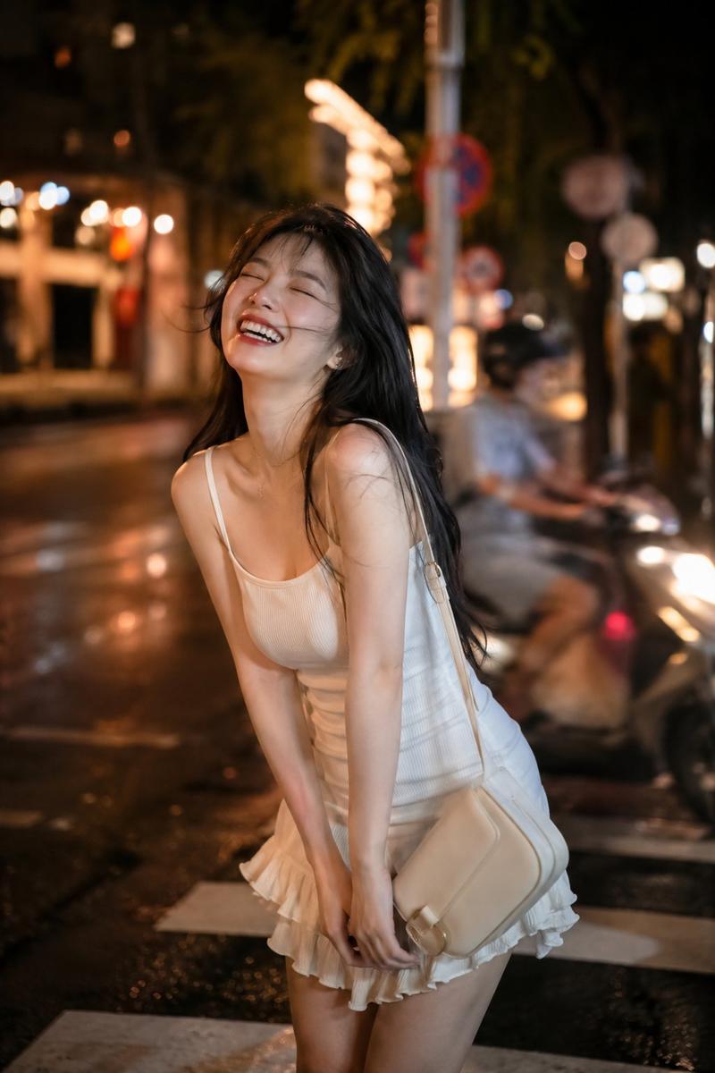一位美丽的年轻东亚女性站在夜晚热闹的街道上，身穿合身的白色细肩带迷你连衣裙，裙摆带有褶边，手提一个奶油色单肩包。她闭着眼睛，头微微后仰，身体前倾...