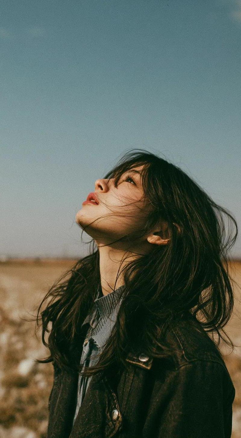 一位美丽的年轻东亚女性独自站在开阔的野花草甸中,头顶是无尽的蓝天,她闭眼或半睁眼向上凝视,仿佛在拥抱微风。长长的自然凌乱黑发自由地披散在脸上,柔...